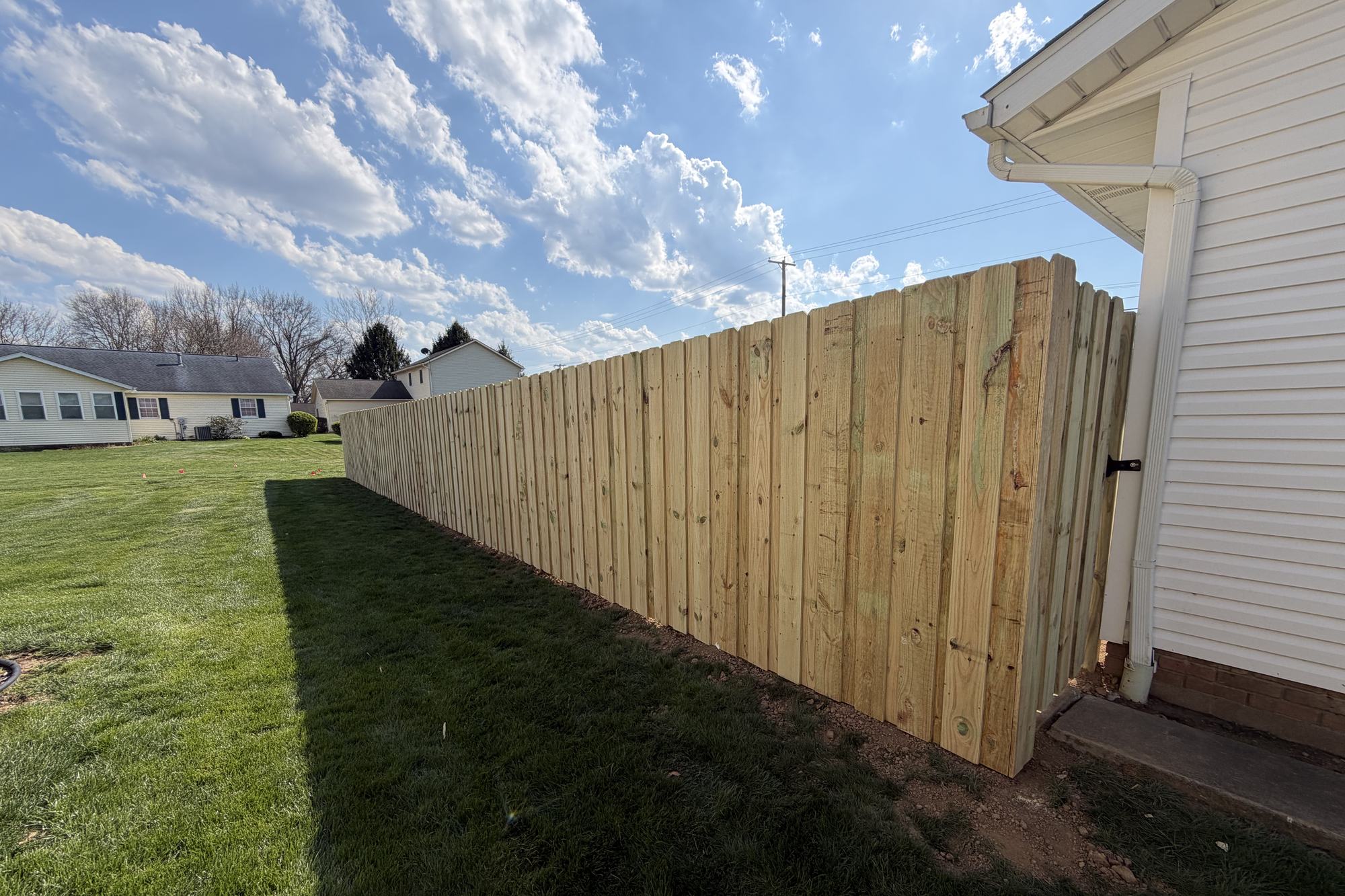 New pressure-treated wood privacy fence running along a backyard beside a white-sided house, installed by Warrior Barriers